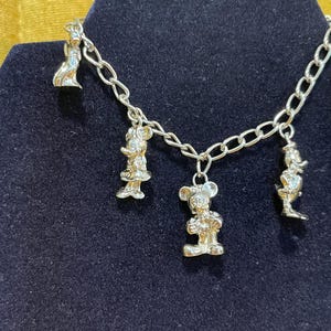 Op de afbeelding: Zilverkleurige bedelarmband met vier cartoonbedels. De bedels zijn bevestigd aan een schakelarmband. De armband wordt gepresenteerd op een donkerblauwe fluwelen ondergrond.