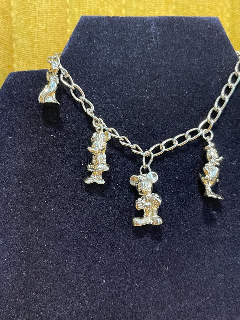 Op de afbeelding: Zilverkleurige bedelarmband met vier cartoonbedels. De bedels zijn bevestigd aan een schakelarmband. De armband wordt gepresenteerd op een donkerblauwe fluwelen ondergrond.