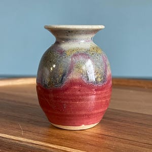 May include: A small ceramic bud vase with a flared rim and a rounded body. The lower portion features a solid red glaze with subtle horizontal ridges, while the upper section transitions into a mottled blend of grey, yellow, and purple tones.