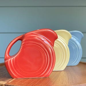 May include: Three ceramic pitchers in red, yellow, and blue. Each pitcher has a handle and a spout. The pitchers are arranged in a row, with the red pitcher in the foreground. The pitchers are on a wooden surface.