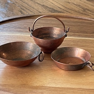May include: Three small, hammered copper vessels with metallic finishes. One vessel features a tall, arched handle, while the other two are shallow bowls, each equipped with a single side ring handle for easy gripping and handling of the items.