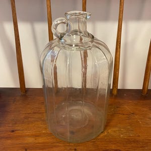 May include: A clear glass jug with a handle and a ribbed design. The jug has a rounded body and a small opening at the top. It is sitting on a wooden surface.