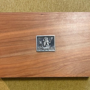 May include: A rectangular wooden box with a decorative metal mermaid emblem. The box is made of reddish-brown wood with a smooth finish. The emblem is a square, detailed with a mermaid figure and wave details.