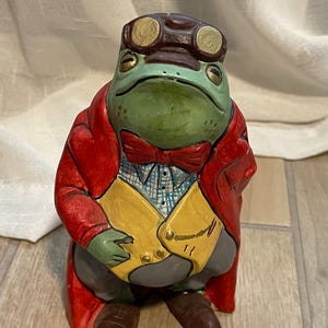 May include: A whimsical ceramic frog figurine, dressed in a red coat, yellow vest, and a bow tie. The frog wears aviator goggles and brown shoes. The figurine has a green face and is posed as if it is standing.