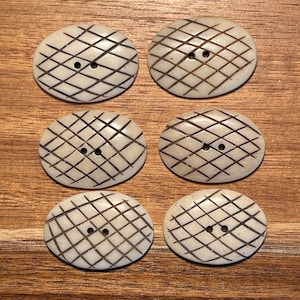 May include: Six oval-shaped off-white buttons arranged in two columns. Each button features a dark brown etched grid pattern across the surface and two small holes positioned in the center for attachment to fabric or other materials.