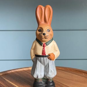 May include: A whimsical, hand-painted Easter bunny figurine. The bunny wears a cream-colored shirt with a red polka-dot tie, gray and white striped pants, and black shoes. The bunny's ears and hands are orange, and it has a green collar.