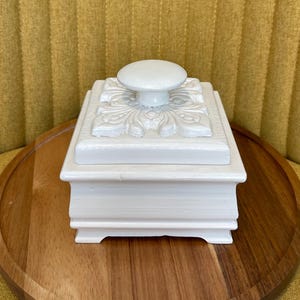 May include: A white, square trinket box with a decorative floral design on the lid. The lid has a round knob for opening. The box is sitting on a wooden tray, with a yellow background.