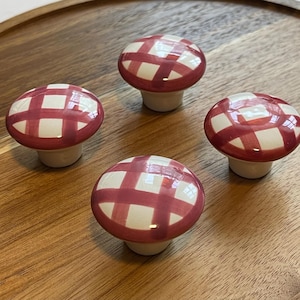 May include: Four mushroom-shaped cabinet knobs with a red and white checkered pattern. The knobs have a glossy finish and are set against a wooden surface. These decorative knobs are ideal for adding a whimsical touch to furniture.