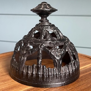 May include: A dark, ornate metal cloche with a decorative finial. The cloche features an openwork design with intricate patterns and a slightly flared base. The overall color is a dark, aged metal finish.
