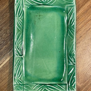 May include: A rectangular ceramic tray with a glossy emerald green glaze and a visible crackle finish. The tray features a raised border decorated with carved, linear leaf-like patterns that frame the smooth, flat central surface area.