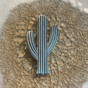 May include: A light blue metal cactus-shaped decorative object. The cactus has vertical lines and a detailed design. The object is set against a textured, cracked, beige background with a white border.