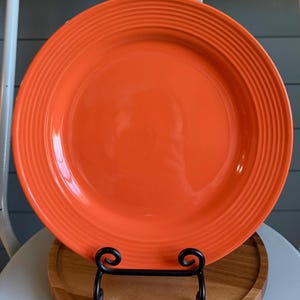 May include: A round, bright orange ceramic dinner plate featuring a series of concentric, raised circular ridges along the wide rim. The plate has a smooth, glossy finish and is displayed upright on a black metal stand.
