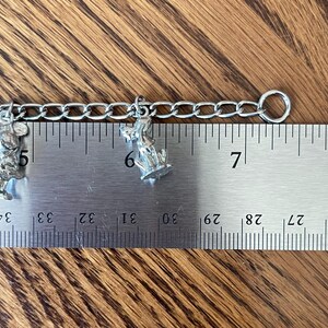 Op de afbeelding: Een zilverkleurige bedelarmband met twee kleine bedels. E&eacute;n bedel is een muis, de andere een hond. De armband wordt gemeten met een liniaal, de ketting is ongeveer 7,6 cm lang.