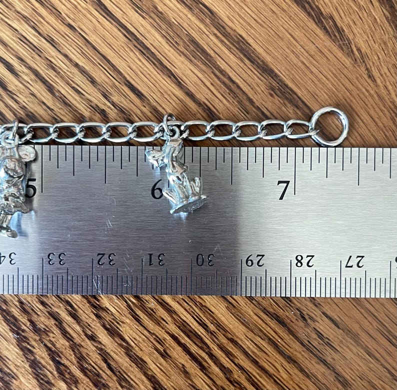 Op de afbeelding: Een zilverkleurige bedelarmband met twee kleine bedels. E&eacute;n bedel is een muis, de andere een hond. De armband wordt gemeten met een liniaal, de ketting is ongeveer 7,6 cm lang.