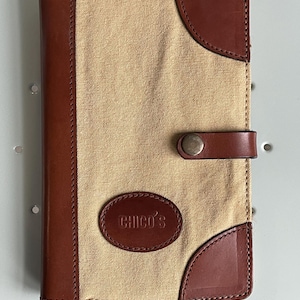 May include: A tan canvas and brown leather wallet with the brand name "Chico's" embossed on a leather patch. The wallet has brown leather trim and a snap closure.
