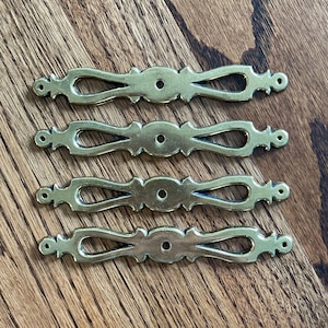 May include: Four brass drawer pulls with an ornate, symmetrical design. Each pull features a central oval shape with a drilled hole, flanked by decorative curves and rounded ends. The pulls are arranged on a wooden surface.