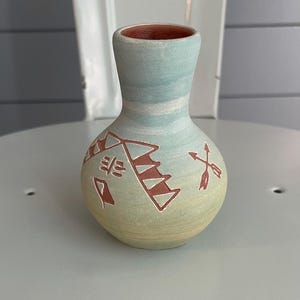 May include: A small, handcrafted ceramic vase with a rounded body and a narrow neck. The vase features a light blue and cream color gradient, with reddish-brown geometric designs and an arrow motif. The interior of the vase is reddish-brown.
