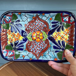 May include: A rectangular ceramic tray featuring a vibrant hand-painted pattern with a central orange flower, intricate brown scrollwork, and yellow grid sections. The design includes blue, green, and red floral motifs on a white background.