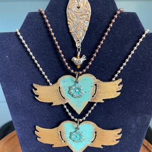 May include: Three necklaces on ball chains. The top pendant is a textured silver-tone heart with a small bird charm. The two lower pendants are identical heart shapes with wings, featuring a turquoise center, gold-tone edges, and a floral accent.