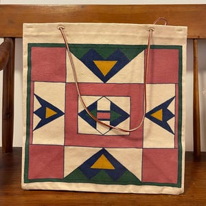 May include: A cream-colored tote bag with a geometric design in pink, green, blue, and yellow. The design features squares, triangles, and diamond shapes. The bag has brown leather handles.