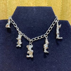 Op de afbeelding: Zilverkleurige bedelarmband met cartoonkarakterbedels. De armband met schakelketting heeft verschillende bungelende bedels van bekende personages. De armband wordt gepresenteerd op een donkerblauwe ondergrond tegen een gouden achtergrond.