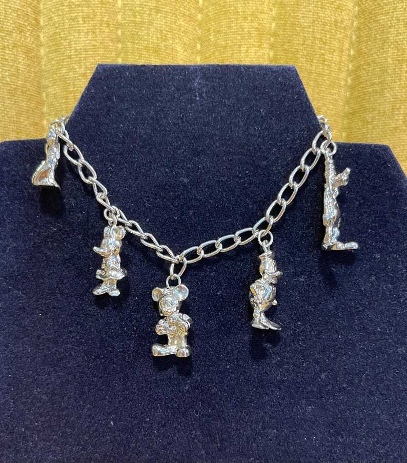 Op de afbeelding: Zilverkleurige bedelarmband met cartoonkarakterbedels. De armband met schakelketting heeft verschillende bungelende bedels van bekende personages. De armband wordt gepresenteerd op een donkerblauwe ondergrond tegen een gouden achtergrond.