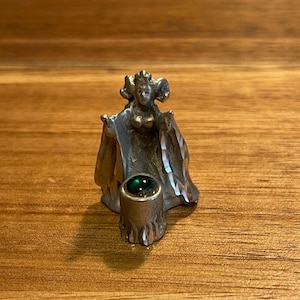 May include: A small, silver-toned figurine of a queen with a flowing gown and crown. The queen holds her arms out to the sides. A cylindrical base with a green gem is at the base of the queen. The figurine is on a wooden surface.