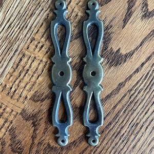 May include: Two antique bronze-colored drawer pulls with an ornate, elongated design. Each pull features a central oval shape with a hole, flanked by decorative elements and a teardrop-shaped opening. The pulls are set against a wooden background.