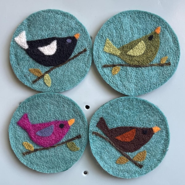 Peut inclure: Un ensemble de quatre dessous de verre circulaires en feutre bleu clair. Chaque dessous de verre présente un oiseau de couleur différente perché sur une branche brune avec de petites feuilles. Les oiseaux sont noir et blanc, vert olive, magenta et brun foncé.