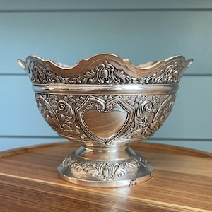 May include: Ornate silver bowl with a scalloped rim and pedestal base. The bowl features detailed floral and shield-shaped designs. The bowl is likely made of silver-plated metal and is displayed on a wooden surface.
