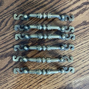 May include: Six antique brass drawer pulls with ornate scrollwork ends and a central decorative element. The pulls are arranged in a parallel pattern on a wooden surface, showcasing their vintage design and potential for furniture restoration.