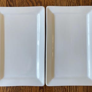 May include: Two identical rectangular white ceramic serving platters with smooth, glossy surfaces and slightly raised edges. These minimalist kitchen dishes feature a clean, solid white finish and a simple, functional design for food presentation.