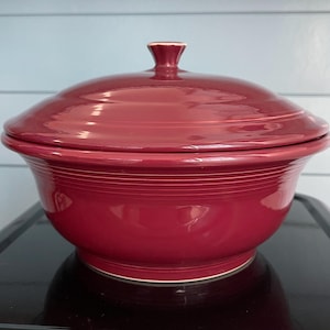 May include: A deep red ceramic serving bowl with a matching lid. The bowl has a wide, rounded shape with a series of concentric rings around the exterior. The lid has a knob on top. The bowl is sitting on a black surface.