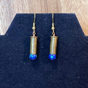 May include: A pair of drop earrings featuring brass bullet casings suspended from gold-toned metal hooks. Each casing is capped at the bottom with a round, vibrant blue bead that displays a speckled, iridescent pattern across its surface.