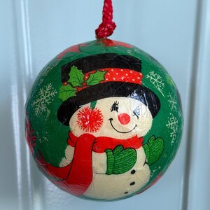 May include: A spherical Christmas ornament with a crackled green finish featuring a snowman wearing a black top hat with holly, a red scarf, and green mittens. The ornament is decorated with white snowflakes and hangs from a red braided cord.