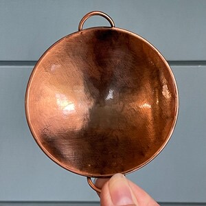 May include: A circular copper bowl with a polished, reflective surface and a smooth, rounded interior. The bowl features two small, semi-circular metal loops attached to the top and bottom edges, providing a symmetrical design for handling.