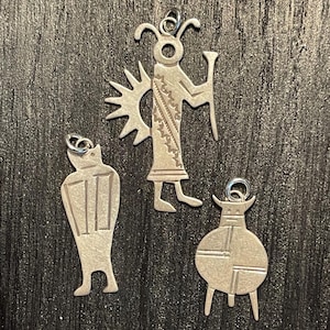 May include: Three silver-toned metal pendants featuring stylized figures. One depicts a flute player with a sunburst, another is a tall rectangular figure with vertical lines, and the third is a round figure with geometric markings and two legs.