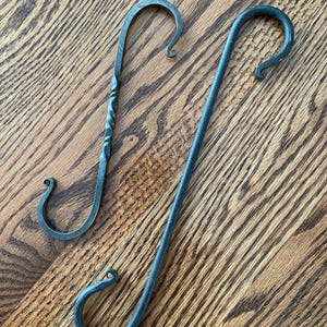 May include: Two dark gray metal S-hooks with curled ends. One hook features a central twisted metal section, while the other hook has a smooth, straight shaft. Both hooks are crafted from forged metal with a matte finish and curved ends.