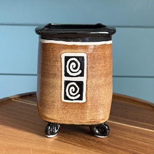 May include: A brown ceramic vase with a black rim and three small feet. The vase features two square designs with white spiral patterns. The vase is a decorative item for the home.