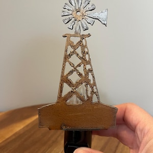 May include: A metal night light featuring a windmill design with a textured bronze-colored tower and a silver-toned fan blade assembly. The structure is mounted on a black electrical plug base, showcasing a rustic finish with visible metal detailing.