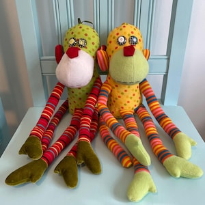 May include: Two handmade plush monkey dolls. One is green with yellow polka dots, the other is yellow with orange polka dots. Both have button eyes, red noses, and striped legs in various colors. The monkeys are sitting on a light blue chair.