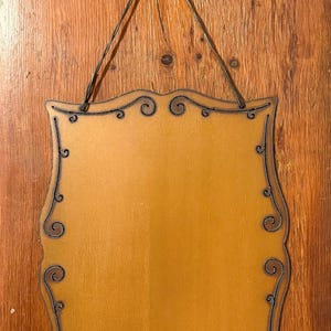 May include: A rectangular wooden sign with a smooth mustard yellow finish and scalloped edges. The perimeter is decorated with dark metal wire detailing in a swirling pattern. A matching dark metal wire loop is attached for hanging.