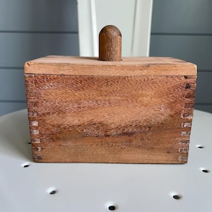 May include: A wooden butter dish with a rectangular box shape and a small, rounded handle on the lid. The wood has a warm, reddish-brown tone and visible grain. The box is constructed with dovetail joints.