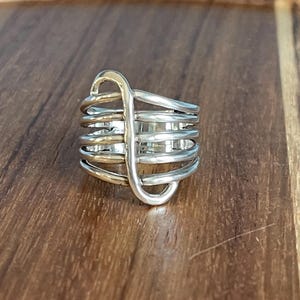 May include: A wide band ring featuring multiple horizontal silver-toned wire loops that are held together by a single vertical wire element. The design creates a layered, cage-like appearance with a polished metallic finish throughout the entire piece.