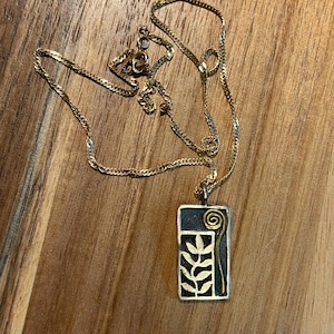 May include: Gold-toned necklace with a rectangular pendant. The pendant features a dark blue background with a gold spiral and a stylized leaf design. The necklace is displayed on a wooden surface.