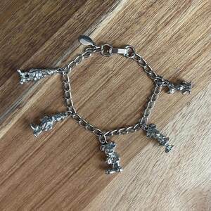 Op de afbeelding: Zilverkleurige bedelarmband met meerdere bedels, waaronder berenfiguren. De armband heeft een schakelontwerp en een sluiting. De armband wordt gepresenteerd op een houten oppervlak. De bedels zijn zilverkleurig.