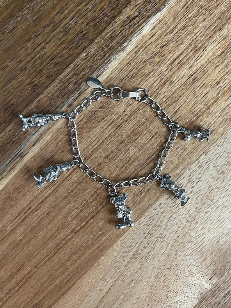 Op de afbeelding: Zilverkleurige bedelarmband met meerdere bedels, waaronder berenfiguren. De armband heeft een schakelontwerp en een sluiting. De armband wordt gepresenteerd op een houten oppervlak. De bedels zijn zilverkleurig.
