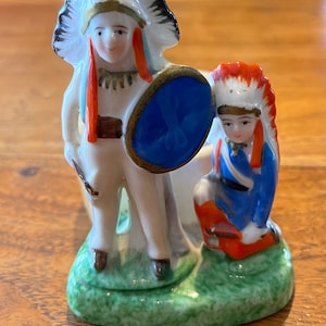 May include: A porcelain figurine of two Native American figures wearing headdresses. The figure on the left is holding a blue shield and the figure on the right is holding a red and white object. The figures are standing on a green base.