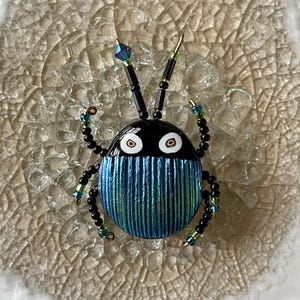 May include: A handcrafted insect-shaped brooch. The brooch features a black head with white eyes and a blue and green striped body. The antennae and legs are made of black beads with accents of green and blue. A unique, artistic accessory.