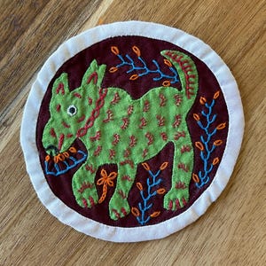 May include: A circular fabric patch featuring a dark maroon background with a central embroidered green dog. The dog has red stitched details and holds a blue flower. Surrounding the dog are blue and orange embroidered floral sprigs.